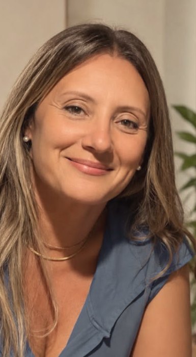 Márcia Campello, psicóloga, de cabelo solto e sorrindo, com blusa azul acinzentada e plantas ao fundo.